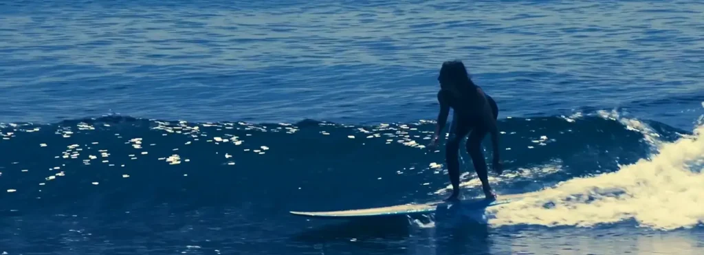 A female surfer in a wetsuit rides a wave, captured in silhouette as she balances on her surfboard with ocean water glistening around her.