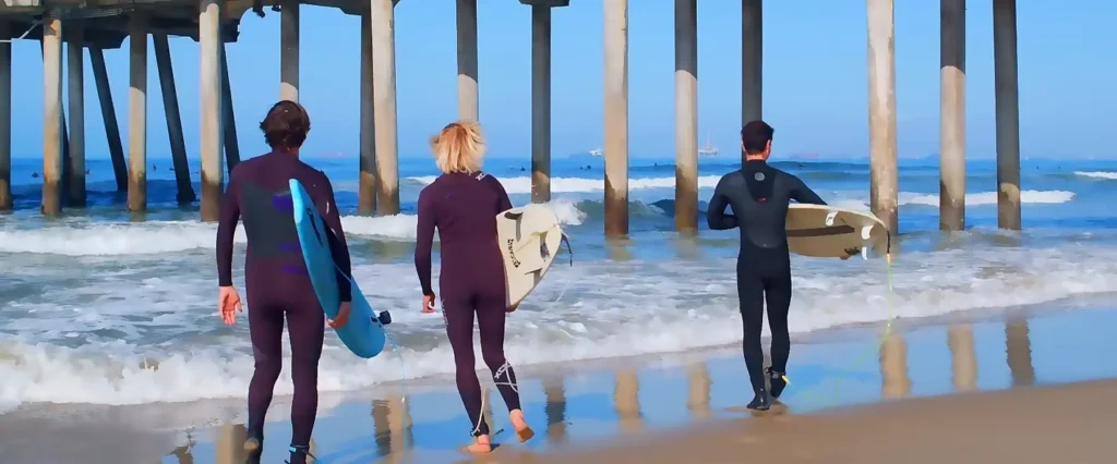 Three surfers — two men and one woman — walk into the ocean carrying their surfboards under a large pier on a bright sunny day.