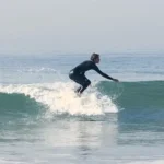 A surfer in a black wetsuit crouches low while riding a breaking wave, carving across the face of the ocean with white water splashing around.