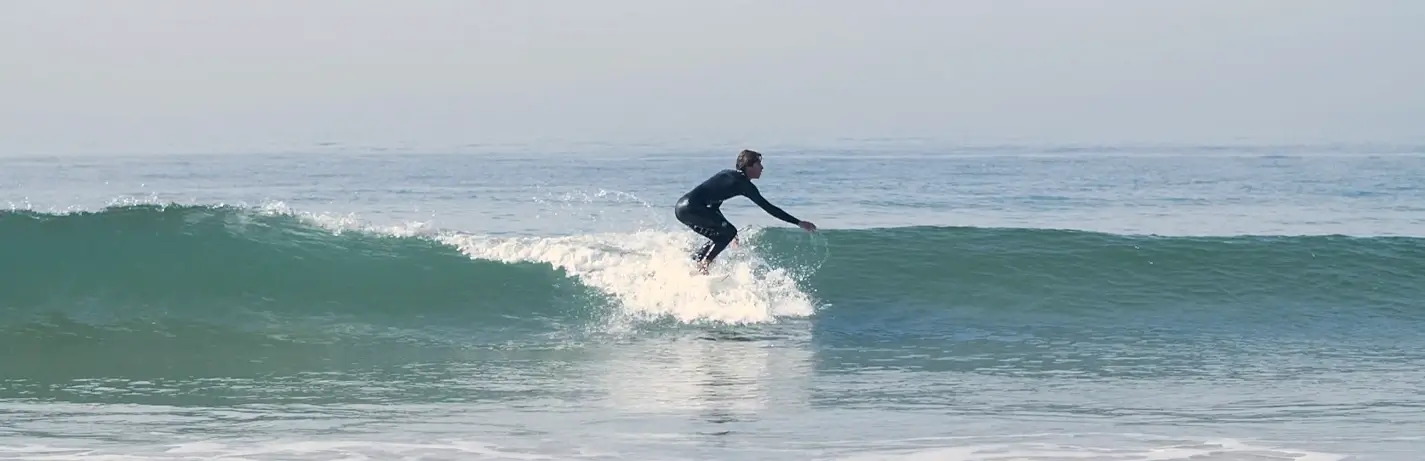 A surfer in a black wetsuit crouches low while riding a breaking wave, carving across the face of the ocean with white water splashing around.