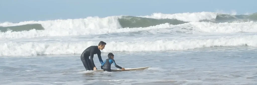 An adult surfer in a full black wetsuit bends over to assist a young child in a wetsuit who is lying on a surfboard in the ocean, with waves breaking in the background.