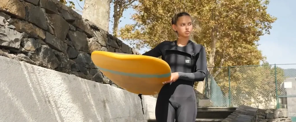 A young woman in a black wetsuit stands outdoors holding a bright yellow surfboard under her arm, looking to the side near a stone wall on a sunny day.
