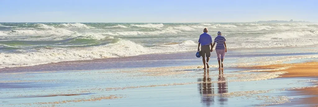 An older couple holding hands and walking along the beach as waves roll in.