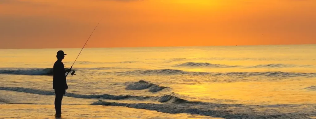 Silhouette of a person fishing from the shoreline against a vivid orange sunset sky.