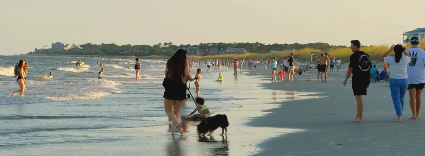 A crowded beach at sunset with people, a dog, and swimmers enjoying the shore.