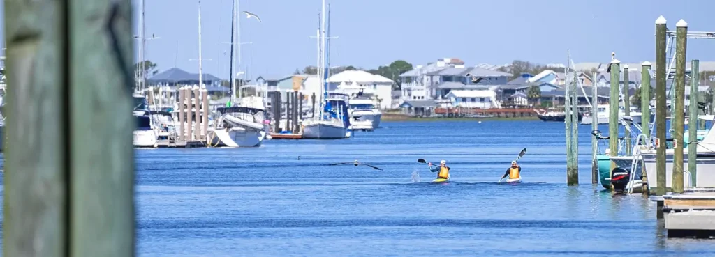 Two kayakers paddling in a marina channel surrounded by docked sailboats and waterfront homes.