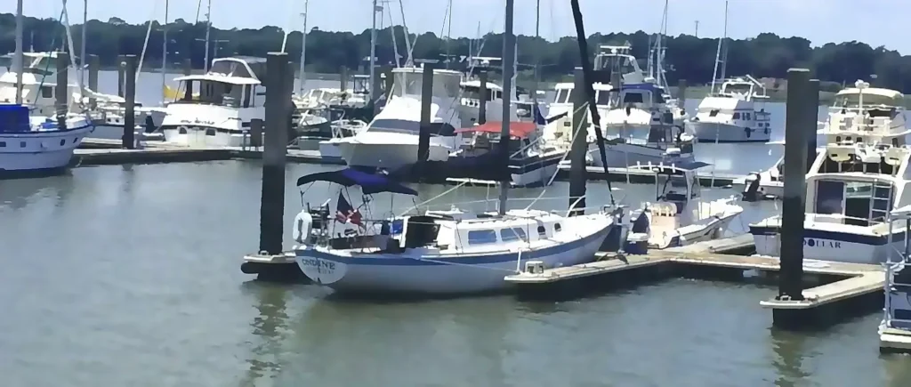 Sunny day at a bustling marina with numerous sailboats and motor yachts docked at wooden piers, including a white and blue sailboat in the foreground with an American flag.