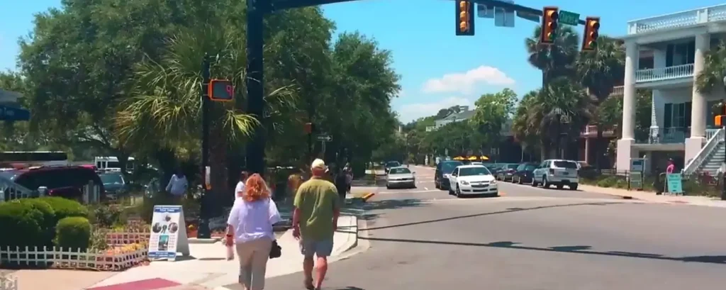 Sunny street scene in historic Beaufort with pedestrians walking along the sidewalk, a woman in a white top and a man in a green shirt in the foreground, palm trees lining the road, traffic lights, parked cars, and a large white colonial-style building with columns on the right under a bright blue sky