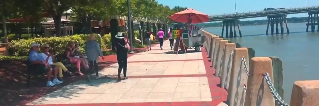 Sunny waterfront boardwalk lined with people walking and sitting on benches, a red umbrella over a vendor cart, concrete bollards with chains, and a large bridge spanning the water in the distance.