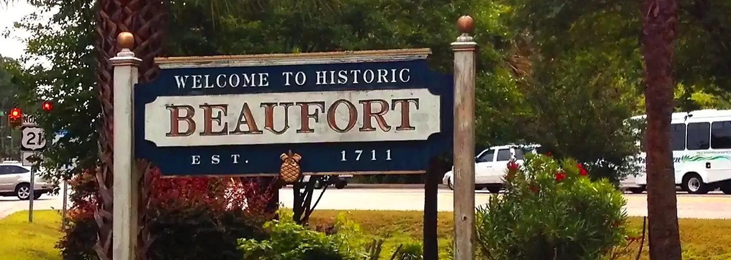 Welcome sign for Historic Beaufort, established 1711, featuring large blue and white lettering with a pineapple emblem, surrounded by palm trees and greenery with vehicles and a road in the background.