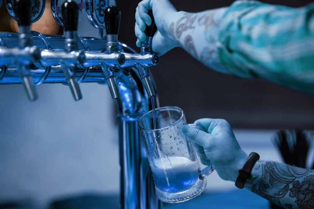 Person with tattooed arms and blue gloves pouring a vibrant blue beverage from a beer tap into a clear glass mug, with white foam forming at the bottom.