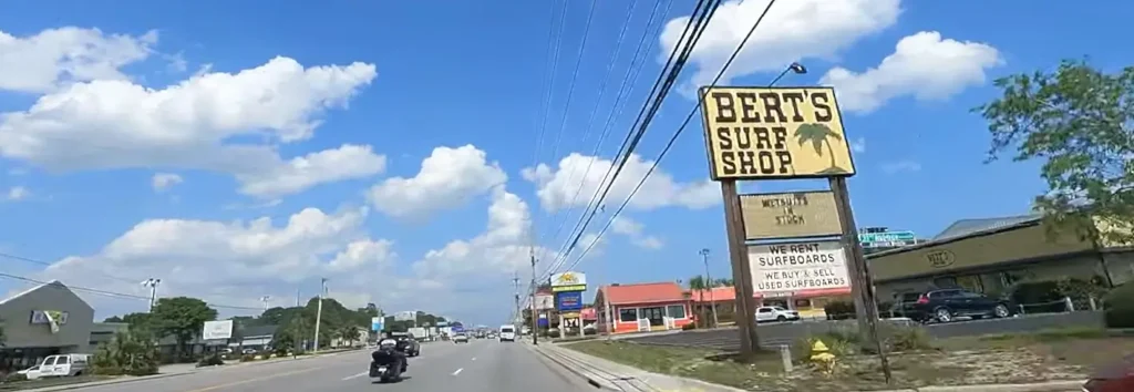 Roadside view in Myrtle Beach featuring Bert’s Surf Shop large wooden sign, with a motorcycle riding down the street on a sunny day.