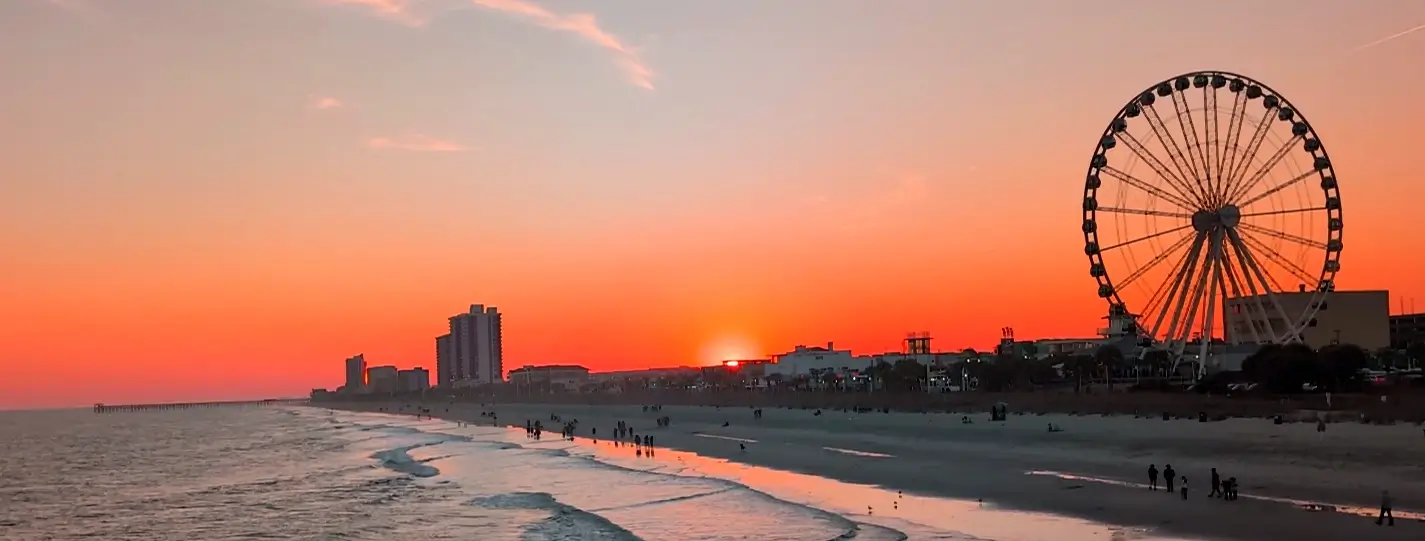 Stunning sunset over Myrtle Beach with a vibrant orange sky, the SkyWheel Ferris wheel on the right, and people walking along the beach as waves gently roll in.