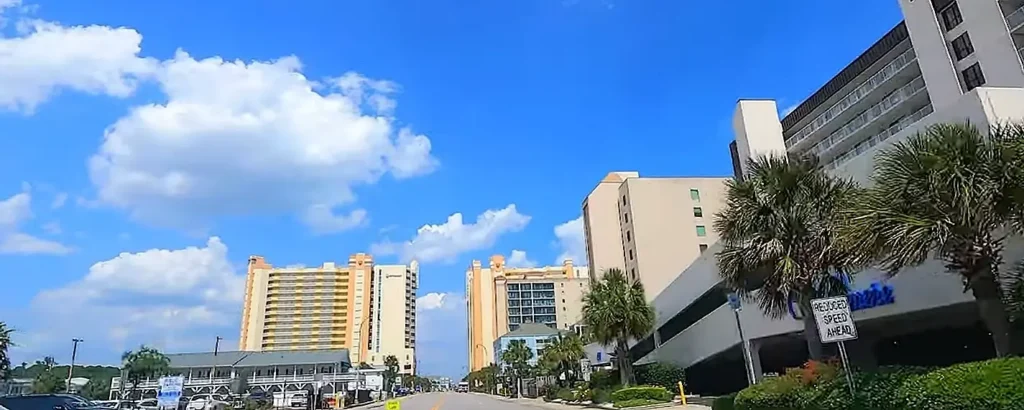 Beachfront high-rise hotels and condos line a bright blue sky day in Myrtle Beach, South Carolina, with palm trees and a parking garage in the foreground.