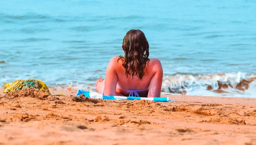 A woman with long brown hair sits on a towel on the sandy beach, facing the ocean with her back to the camera as gentle waves roll in.