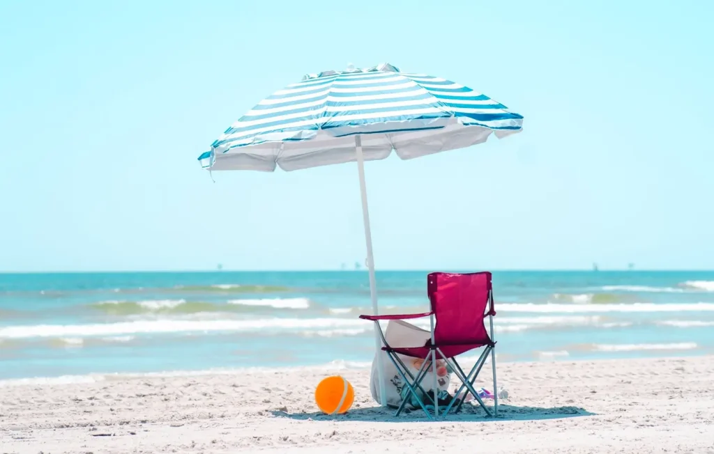 A blue and white striped beach umbrella shades a red folding chair on the white sand, with an orange beach ball nearby and calm ocean waves in the background.