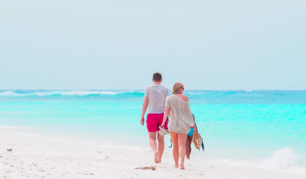 A couple walks hand-in-hand barefoot along the shoreline on a white sandy beach, with turquoise ocean waves and a clear sky stretching into the distance.