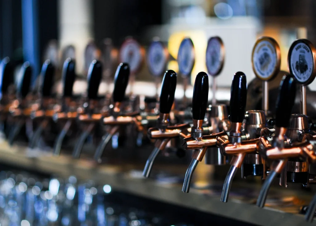 Close-up of multiple beer taps with black handles and copper fixtures lined up along a bar, with blurred background lights and glassware.