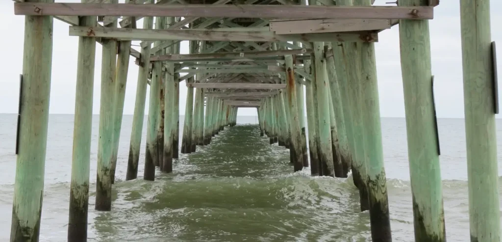 Underneath a long pier at the beach, showing rows of weathered wooden pillars with waves rolling through the structure.