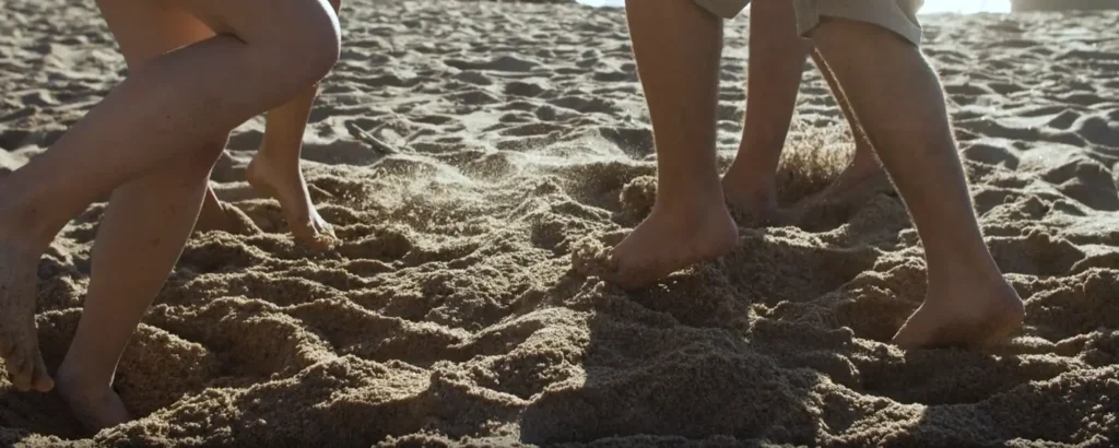 Close-up of several people’s bare feet walking through soft, sunlit beach sand, capturing a carefree summer moment.
