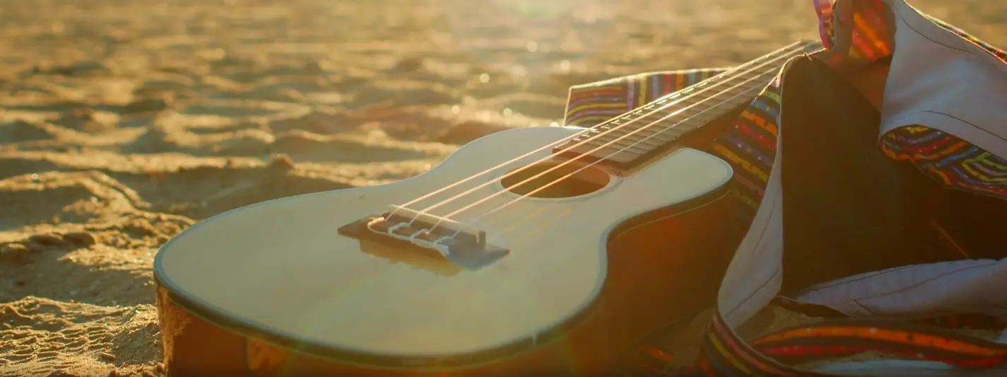 A wooden ukulele resting on the golden sand at sunset, with a colorful woven bag nearby and warm sunlight creating a peaceful beach scene.