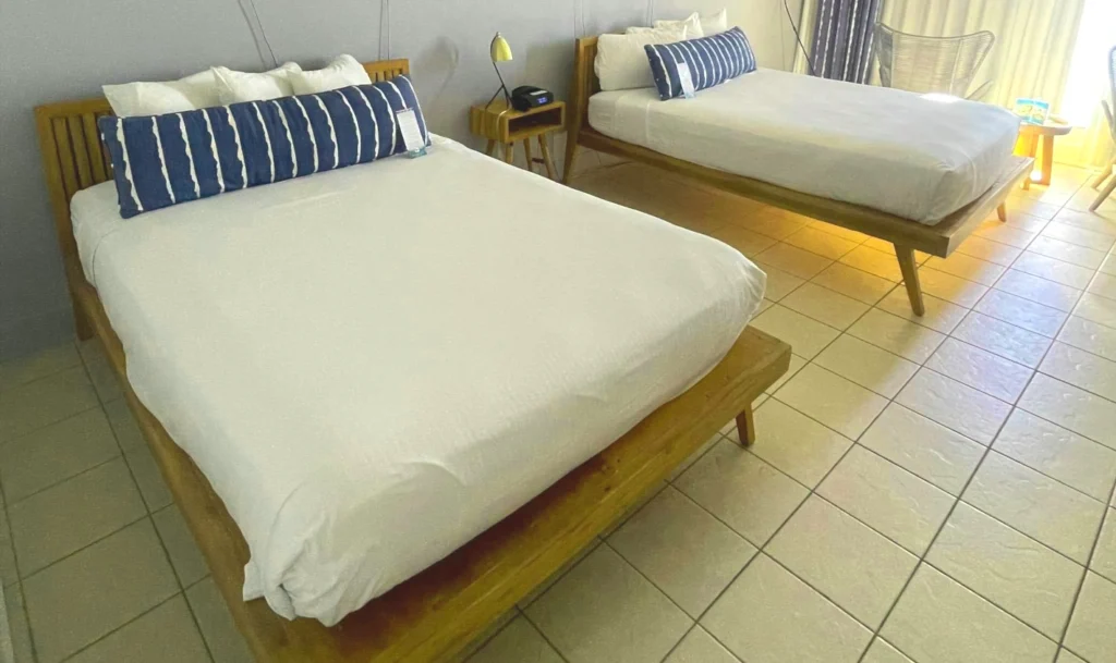 Hotel room with two wooden platform beds featuring white linens and blue-and-white striped accent pillows. Light-colored tiled floor, wooden nightstands, and large windows with curtains in the background