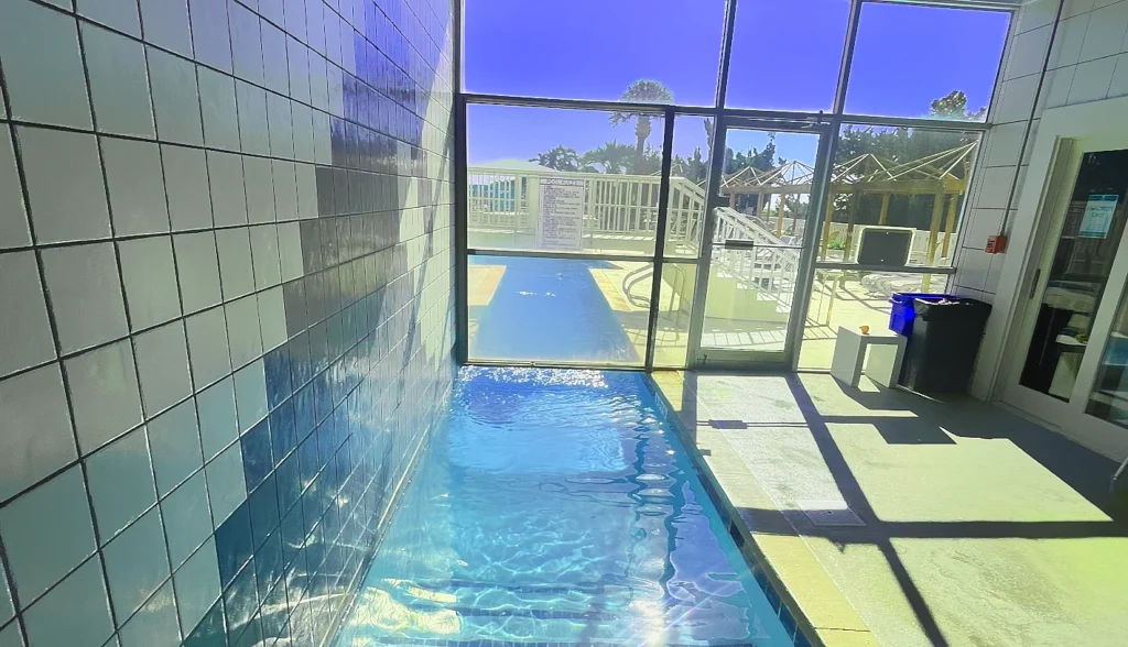Indoor lap pool with bright blue water, white tiled walls, and large glass doors opening to a sunny outdoor pool area with palm trees and clear blue sky. Bright, modern pool enclosure with natural light flooding in.