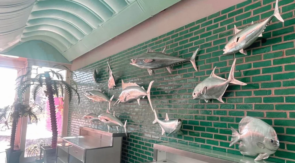 Green brick wall decorated with multiple metallic silver fish sculptures of various sizes and species. A potted palm plant stands to the left near large windows, with stainless steel counters below the display. Nautical/seafood-themed interior.