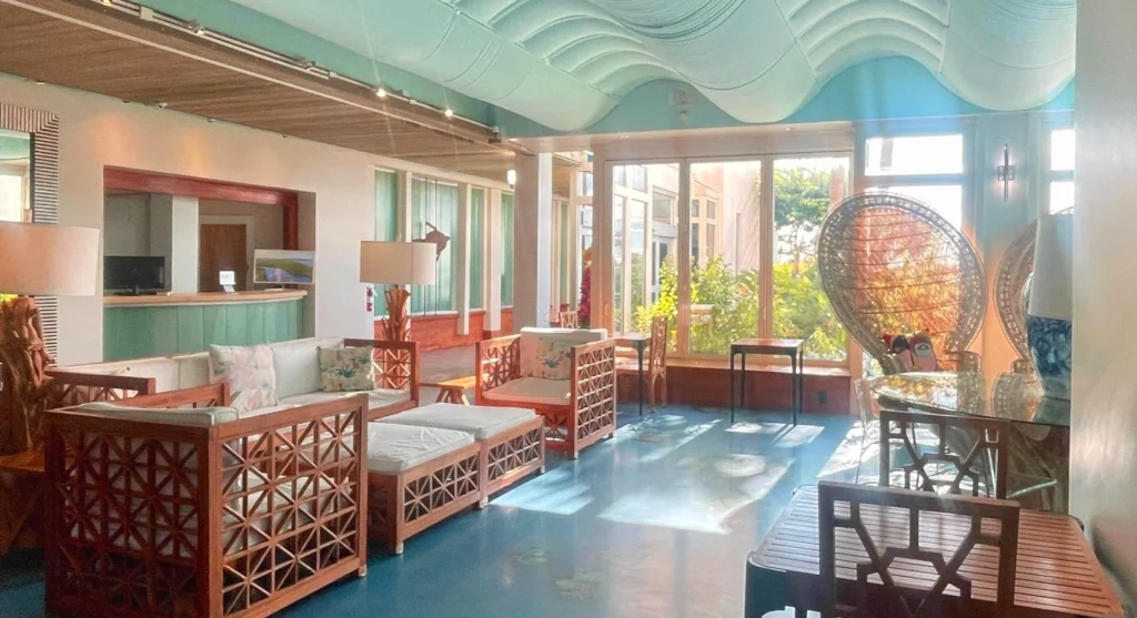 Spacious resort lobby lounge with turquoise wavy ceiling, wooden lattice sofas and armchairs with white cushions, large windows letting in natural light, peacock wicker chairs, and a bar area in the background. Bright, tropical interior with blue floor.
