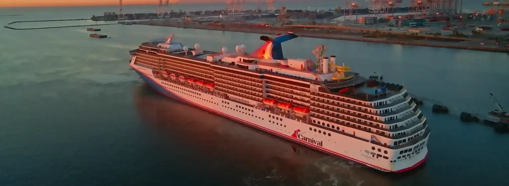 Aerial view of a large Carnival cruise ship (with the distinctive blue-and-red funnel) docked or departing a port at sunset. The massive vessel is illuminated with warm lights, surrounded by harbor infrastructure, cranes, and calm water reflecting the orange sky.