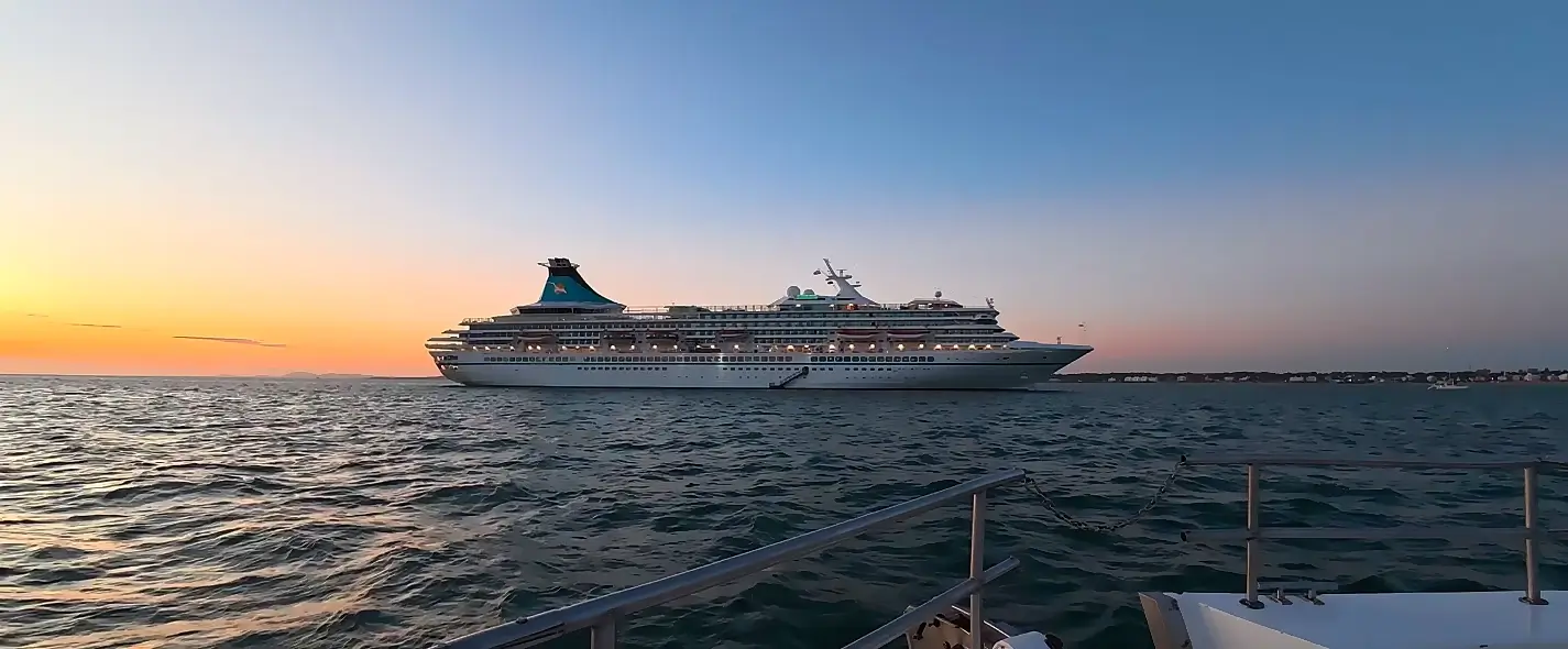 A large white cruise ship sailing on calm water during a golden sunset. The vessel is viewed from another boat (railing visible in foreground), with soft orange and blue twilight colors in the sky and a distant shoreline on the horizon.