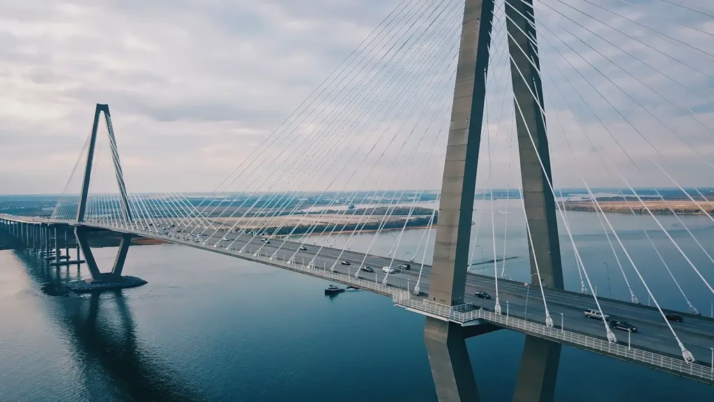 Aerial view of the impressive Arthur Ravenel Jr. Bridge, a modern cable-stayed bridge spanning a wide river. Cars are visible driving across the multi-lane roadway under a partly cloudy sky, with marshland and water stretching into the distance.