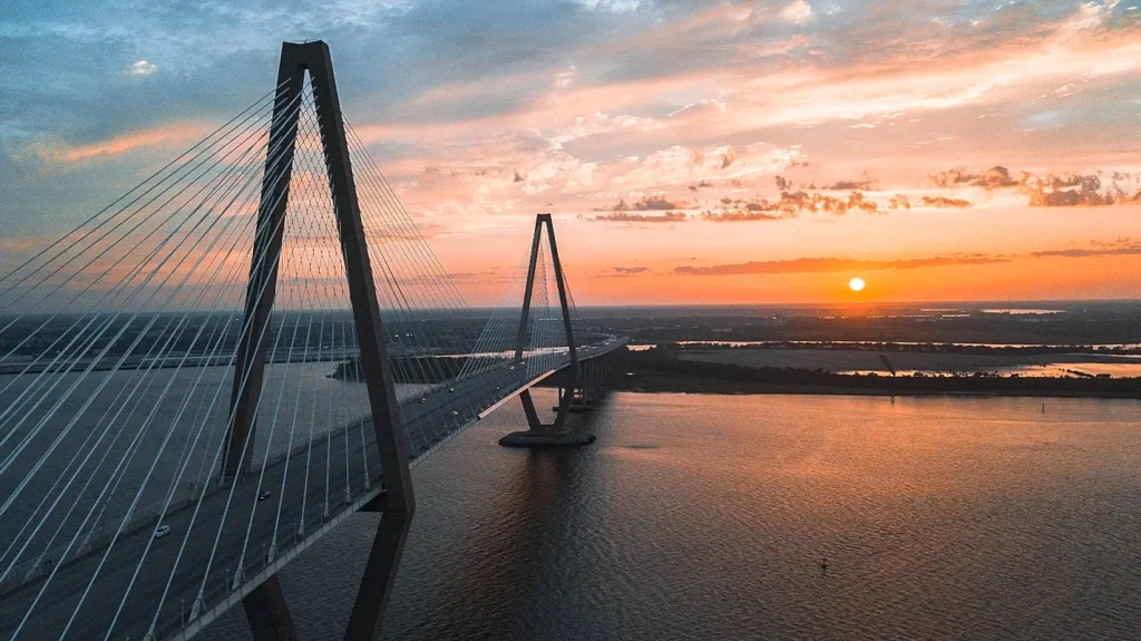 Stunning aerial shot of the Ravenel Bridge at sunset, with dramatic orange and pink clouds reflecting on the calm water below. The bridge’s tall cable towers are silhouetted against the vibrant sky, with vehicles crossing the span and marshland visible in the background.