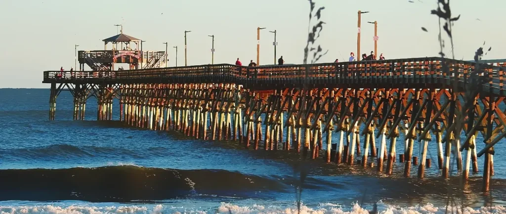 Long wooden pier stretching over the ocean at Cherry Grove Beach, South Carolina, with people fishing and walking along it, waves crashing below under a bright sky.