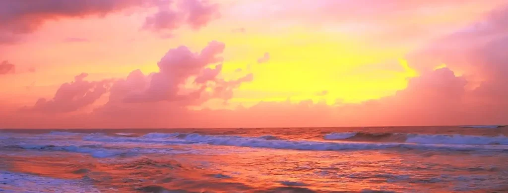 Vibrant sunset over the ocean, with a dramatic sky filled with glowing pink, orange, and yellow clouds. The bright sun sits low on the horizon, casting warm reflections across the water while gentle waves with white foam roll toward the shore.