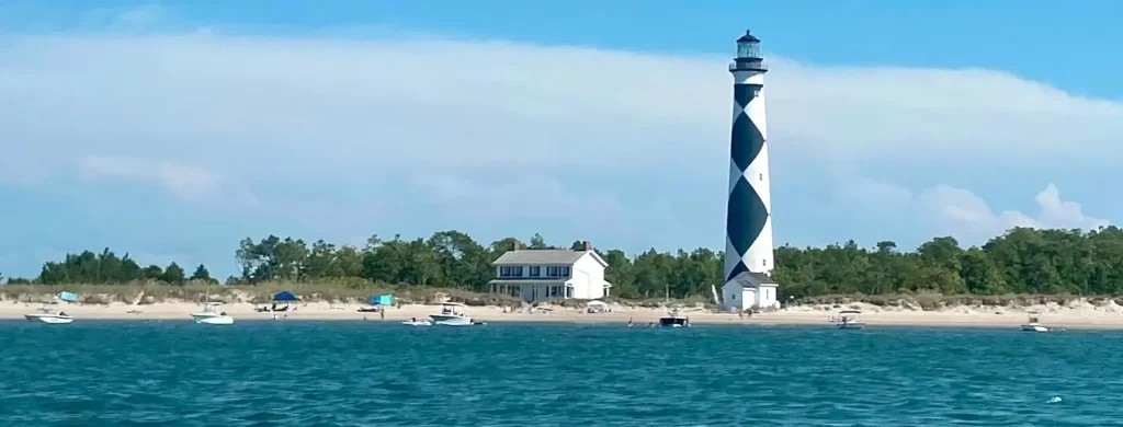 Iconic black-and-white diamond-patterned lighthouse standing tall on a sandy beach next to a white keeper’s house. Several boats are anchored in the turquoise water in the foreground, with people visible on the shore and trees lining the background under a bright blue sky.
