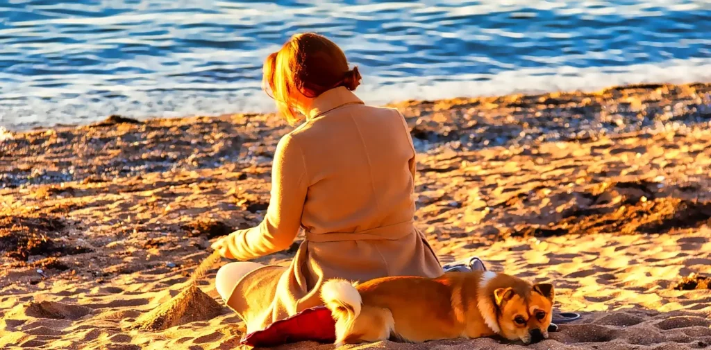A woman with reddish-blonde hair in a beige coat sitting on the beach at sunset, facing the ocean, with a small corgi-type dog lying peacefully beside her.