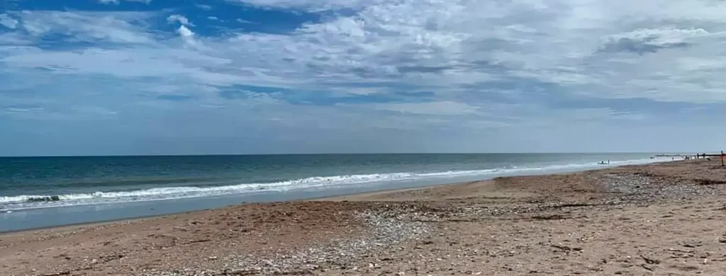 Wide sandy beach with gentle waves rolling onto the shore under a partly cloudy sky. The ocean stretches to the horizon, with a few people visible in the distance along the coastline.