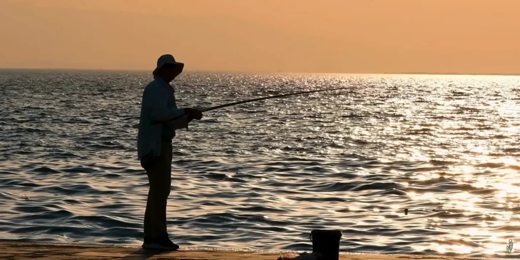 Silhouette of a fisherman wearing a hat and light-colored clothing, standing on a pier or shoreline at sunset. He holds a long fishing rod extended over the golden, rippling sea as the low sun creates a bright reflection on the water. A small bucket sits on the pier nearby.