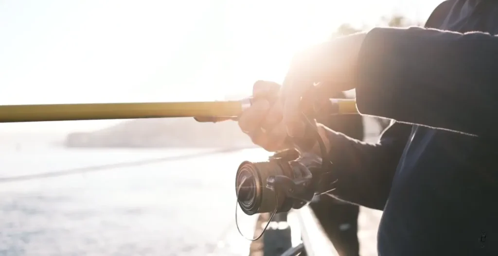 Close-up of a person’s hands gripping a yellow fishing rod and spinning reel, with warm golden sunlight creating a strong lens flare. The background shows blurred water and distant shoreline, suggesting a bright, sunny day by the sea.