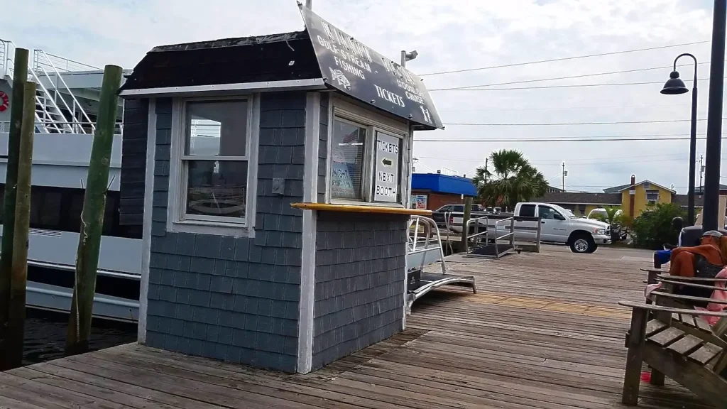 Wooden dock ticket booth for Murray’s Gulf Stream Fishing Cruises. The small blue-gray shingled booth has signs for tickets and 'Next Booth'. A large white tour boat is docked behind it, with people relaxing on benches along the boardwalk on a sunny day.