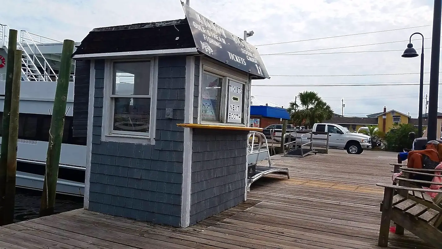 Wooden dock ticket booth for Murray’s Gulf Stream Fishing Cruises. The small blue-gray shingled booth has signs for tickets and 'Next Booth'. A large white tour boat is docked behind it, with people relaxing on benches along the boardwalk on a sunny day.