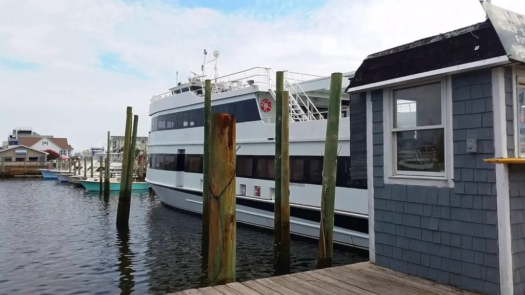 A large white multi-deck passenger boat docked at a wooden pier on a calm day. The vessel is moored next to weathered wooden pilings, with a small gray shingled ticket booth in the foreground and other boats and coastal buildings visible in the background.