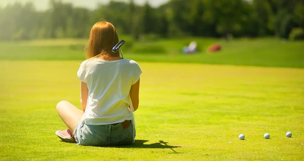 Rear view of a woman with long red hair sitting cross-legged on a sunny golf course, with a golf club resting on her shoulder and three golf balls on the grass nearby.