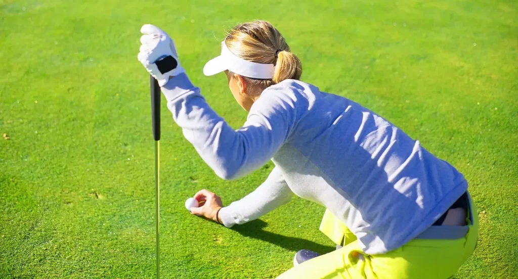 A woman in a blue long-sleeve shirt and neon yellow pants kneels on a golf green, lining up a putt with her putter while wearing a white visor and golf glove.