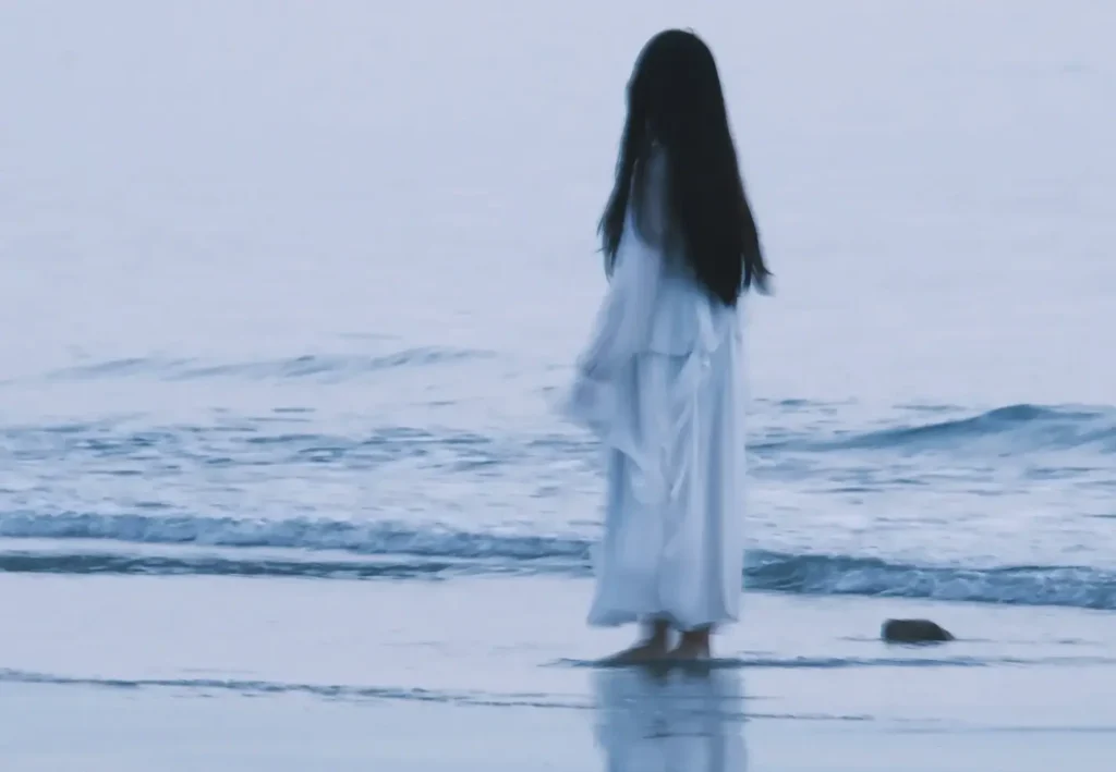 A woman with long dark hair wearing a flowing white dress stands barefoot at the edge of the ocean, looking out at the waves.