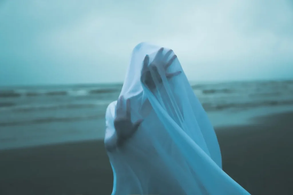 A surreal figure shrouded in a flowing white veil on the beach, with hands pressing against the translucent fabric.