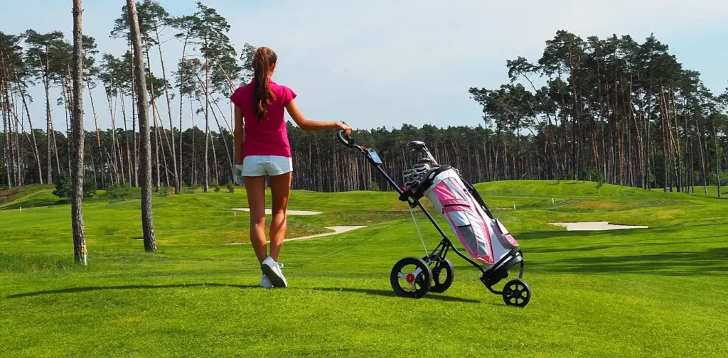 A woman in a pink shirt and white shorts walks across a lush golf course pulling a pink and black golf push cart, with tall pine trees in the background.