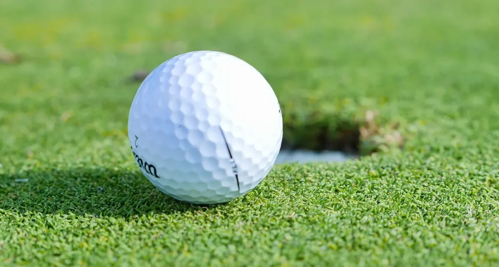 Close-up of a white Wilson golf ball resting on the edge of a golf hole on a bright green putting surface.