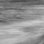 Long exposure black and white photograph of a ghostly blurred figure standing on a sandy beach beside a turbulent sea.